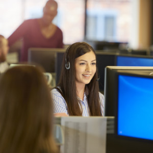 a female customer service representative sits at her computer terminal in a call centre chatting happily over her headset to a customer. In the background and foreground colleagues are seen sitting at their desks .